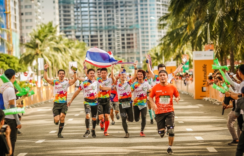 Group of marathon runners in colorful jerseys celebrating with a flag as they cross the finish line past a cheering crowd
