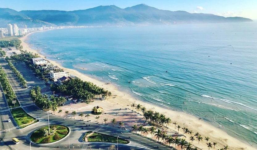 Aerial view of My Khe Beach in Da Nang with long sandy shore, the coastal road and roundabout, and mountains beyond