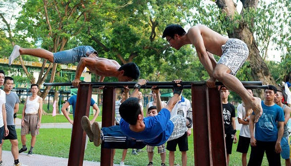 Young men doing calisthenics on outdoor parallel bars in a park while a crowd watches them train