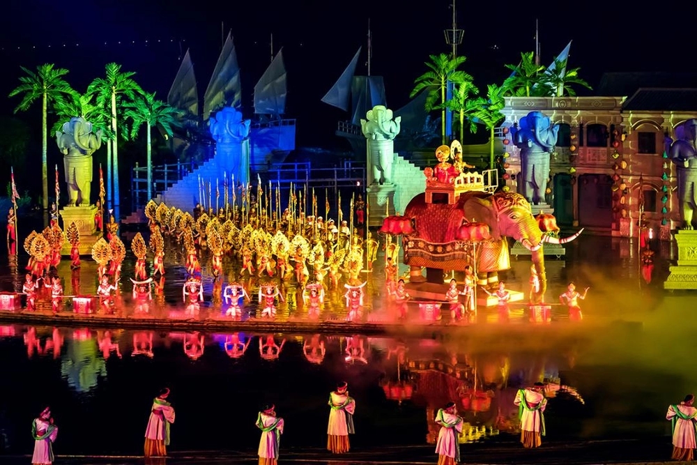 Colorful nighttime outdoor stage show with costumed dancers, a lit elephant float and reflections on water