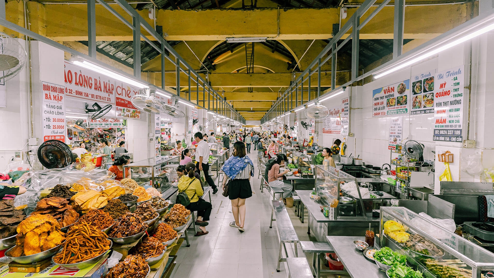 Busy indoor market aisle lined with food stalls displaying local dishes as shoppers walk through Con Market