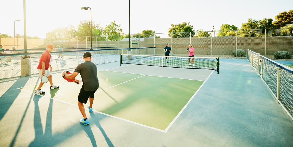 Four adults playing doubles pickleball with paddles on an outdoor court at sunset surrounded by green fencing