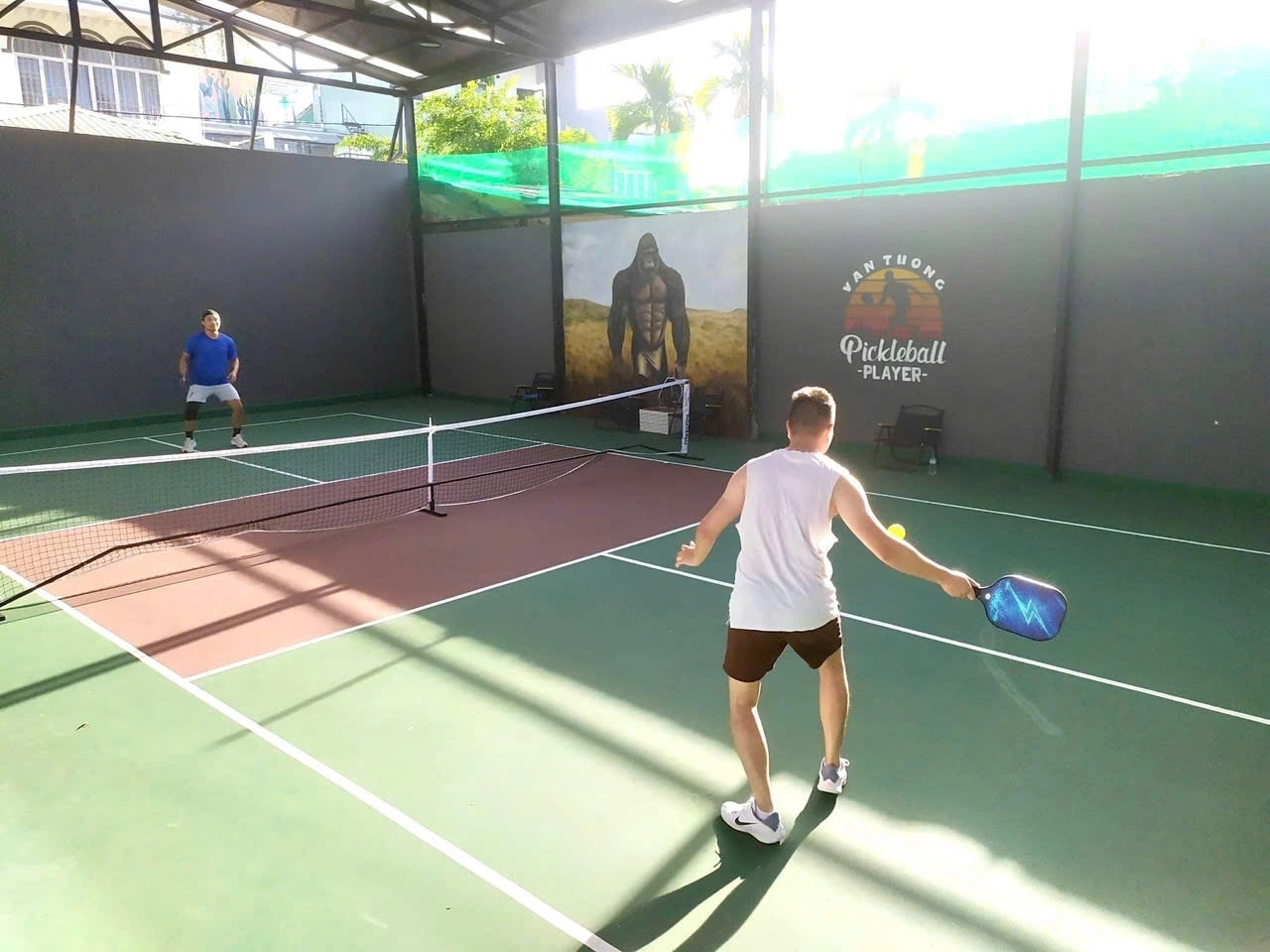 Two men playing pickleball on a covered Van Tuong court with a gorilla mural and player logo on the wall