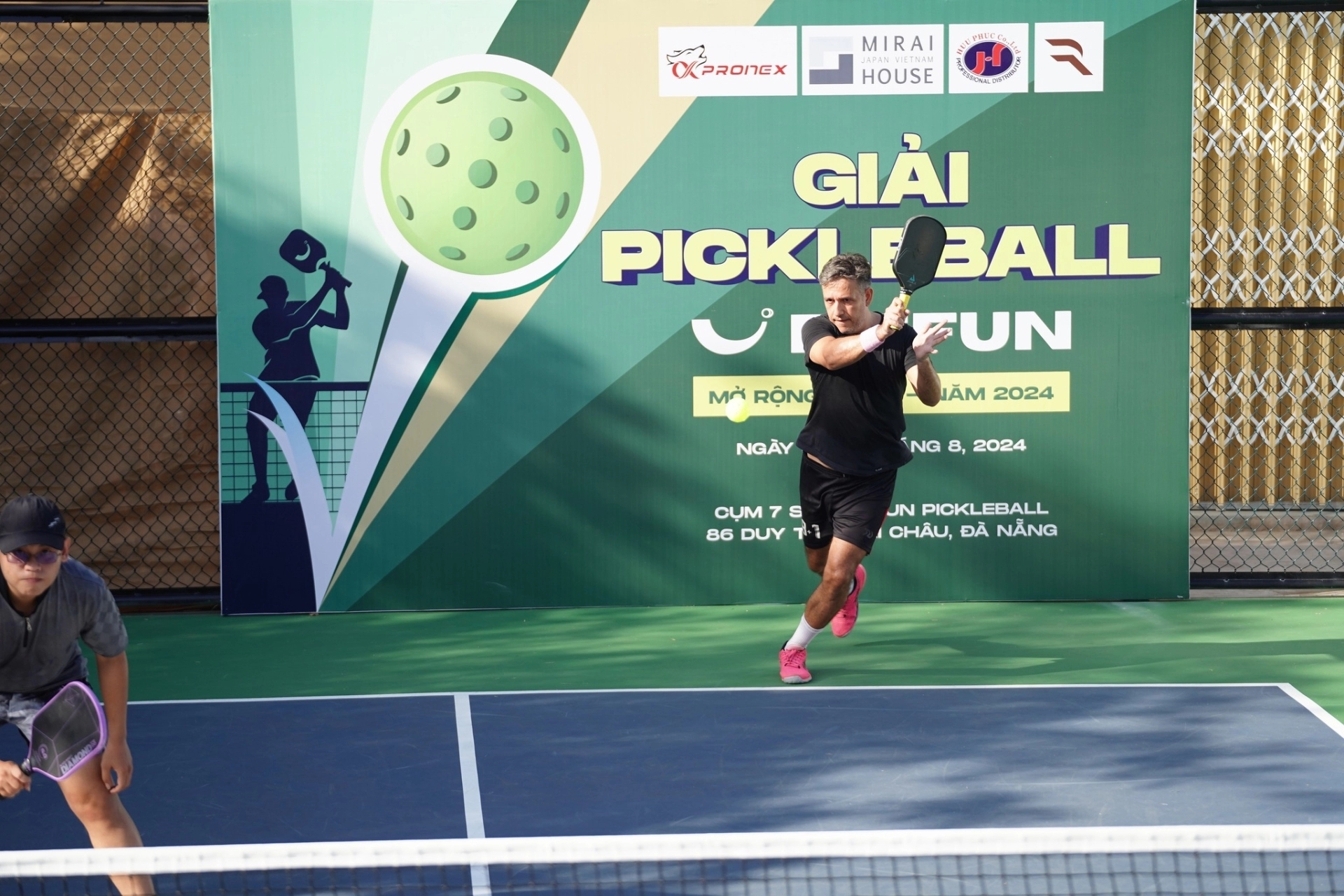 Man swinging a paddle during a pickleball tournament match in front of a green Fit Fun event banner