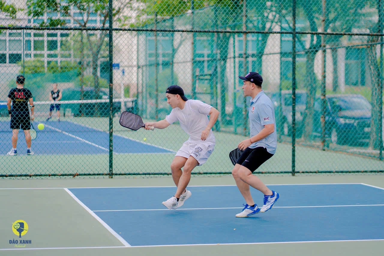 Two young men in light shirts playing pickleball doubles on an outdoor blue court behind chain-link fencing