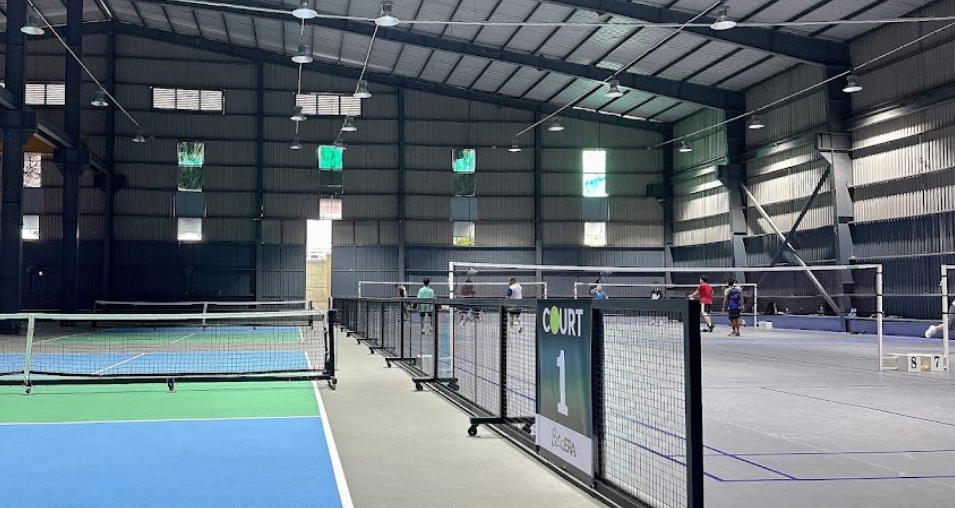 Indoor warehouse-style sports hall with several pickleball courts and a Court 1 sign under a high metal roof