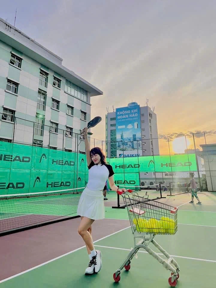 Woman in white holding a paddle and posing with a cart of balls on a rooftop court at sunset with green fencing