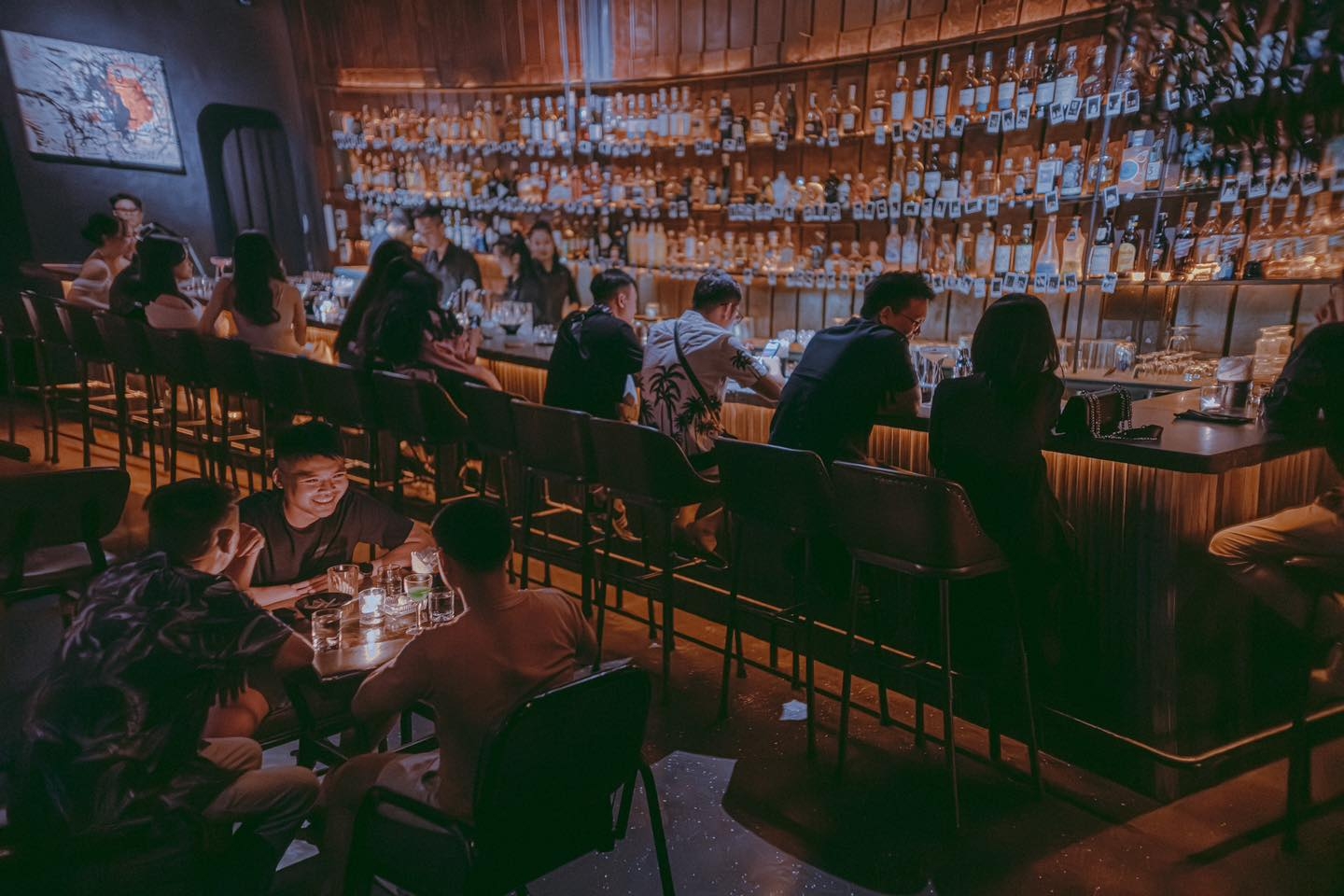 Cozy cocktail bar with a curved wall of bottles, patrons on stools and seated at low candlelit tables