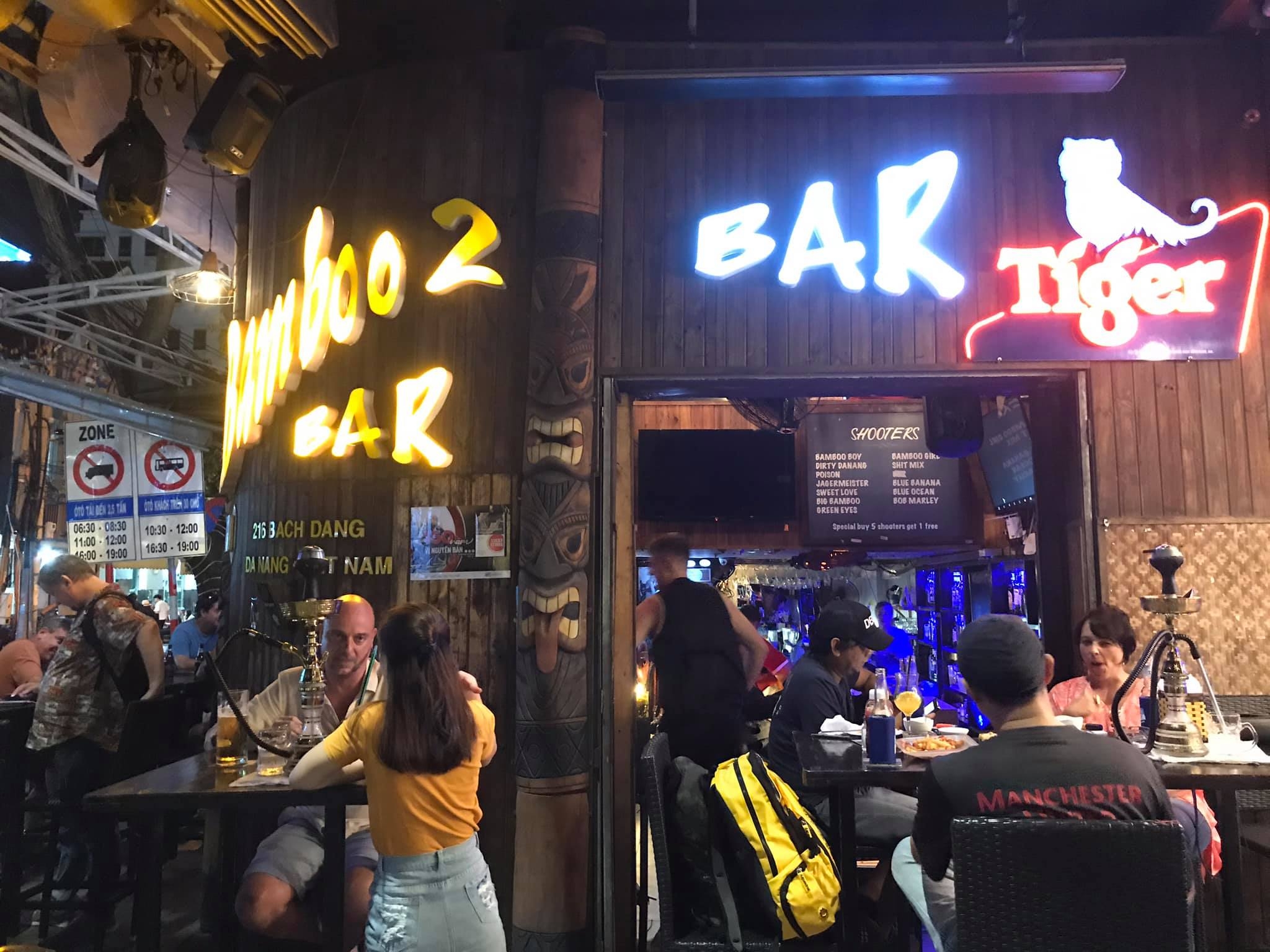 Bamboo 2 Bar street frontage with glowing neon signs, a Tiger beer sign, wooden totem and guests