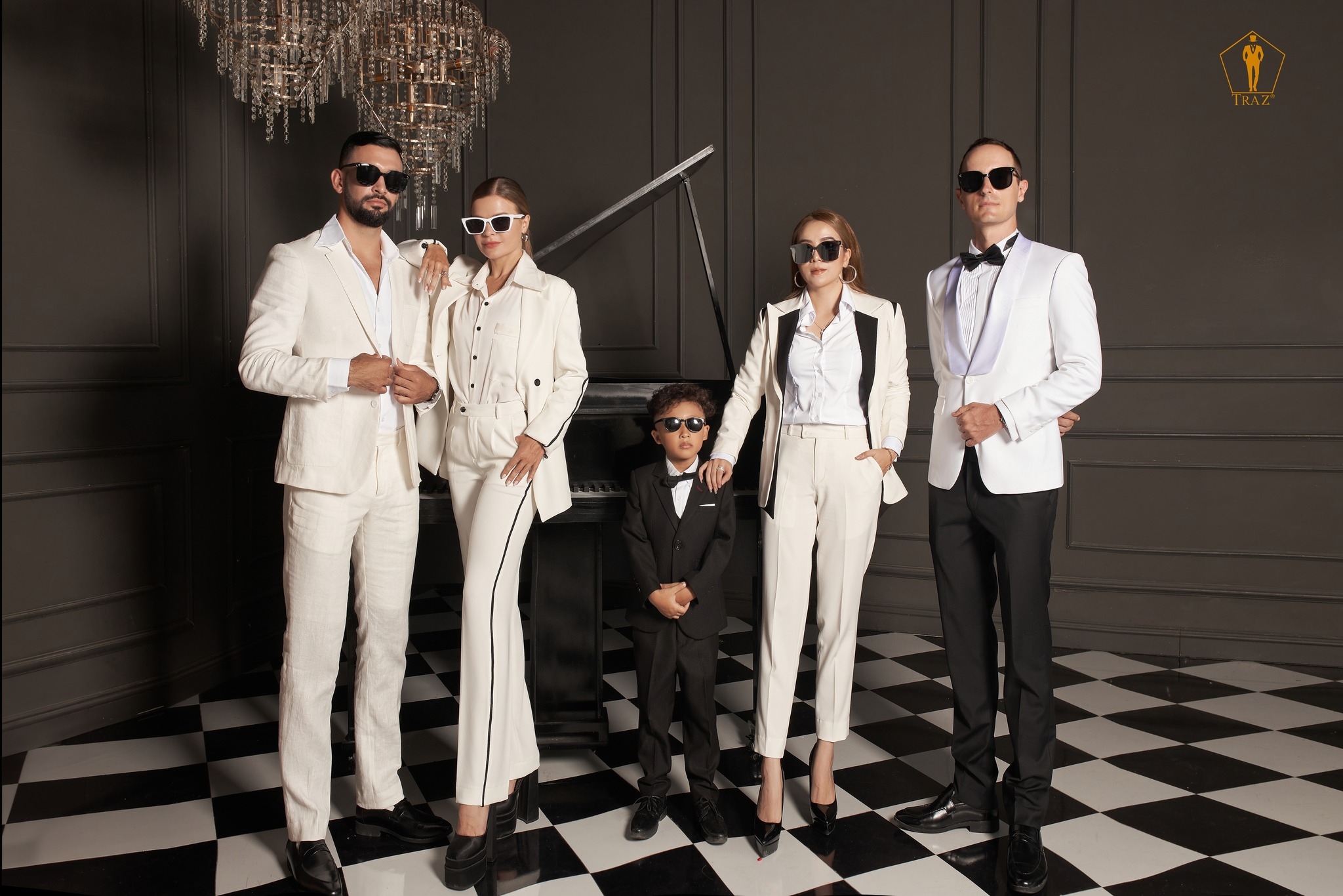 Family of five in cream and black formal suits and tuxedos posing with sunglasses by a grand piano in an elegant room