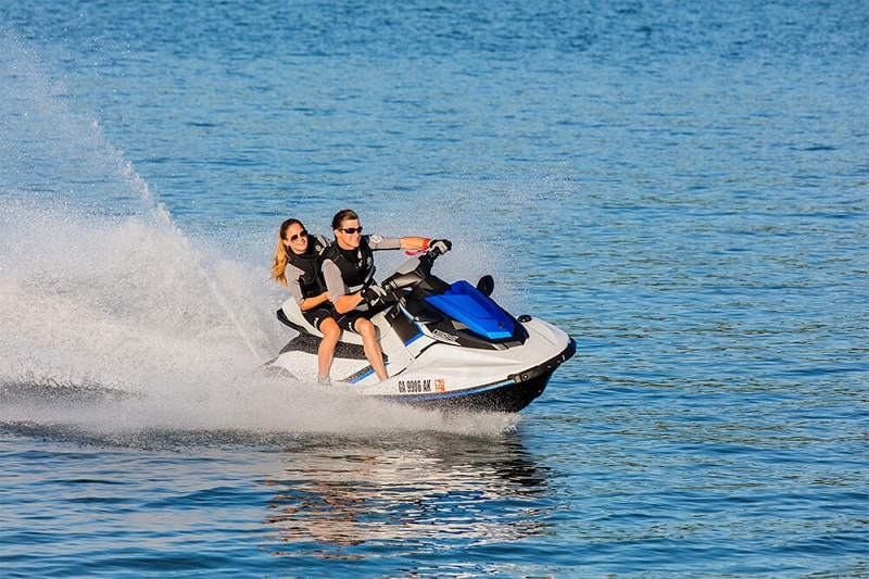 Two people in life jackets riding a white and blue jet ski fast across blue water, spraying a wake behind them