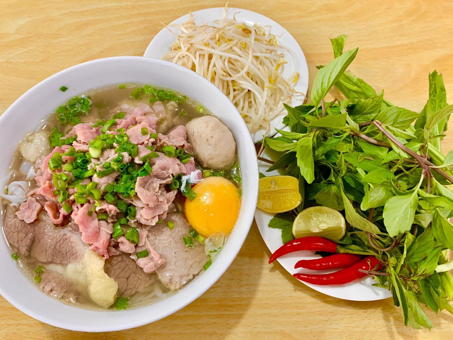 Beef pho bowl with sliced beef, beef ball and raw egg beside a plate of bean sprouts, herbs, lime and chili