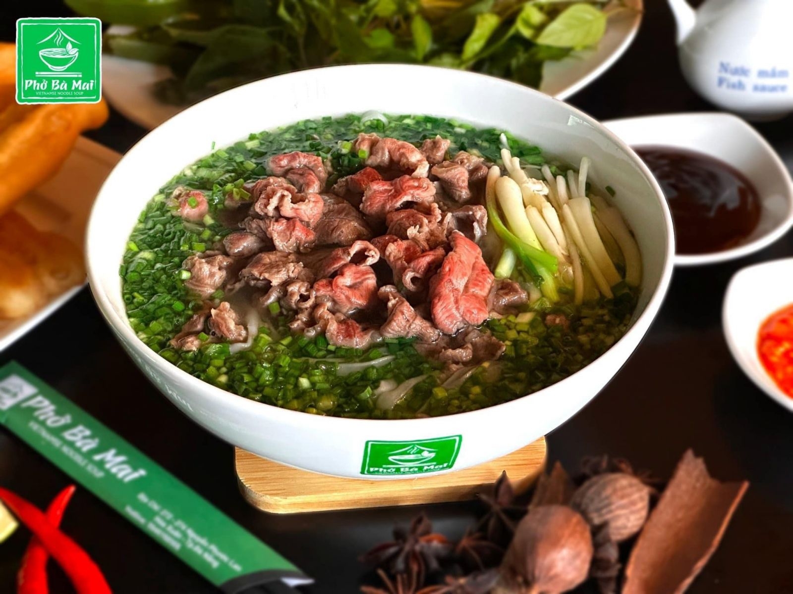 Pho Ba Mai branded bowl of beef pho loaded with chopped chives, beside star anise and cinnamon spices