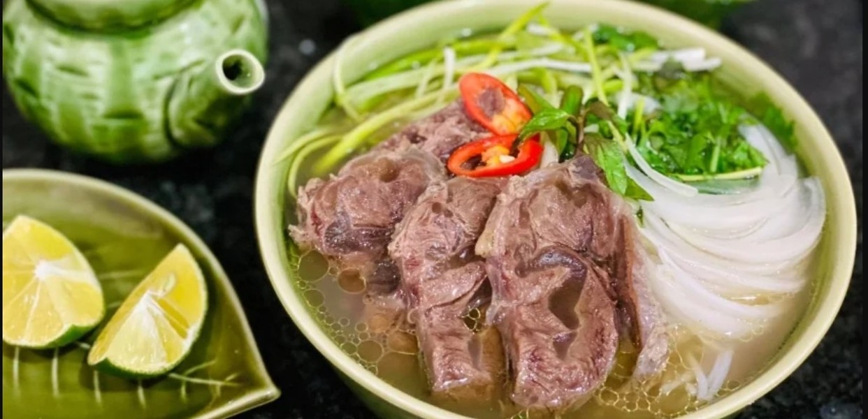 Green bowl of beef pho with rice noodles, red chili and herbs, a green teapot and lime wedges nearby