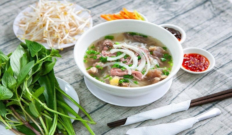Bowl of beef pho with onion and herbs served with bean sprouts, fresh herbs, chili sauce and chopsticks