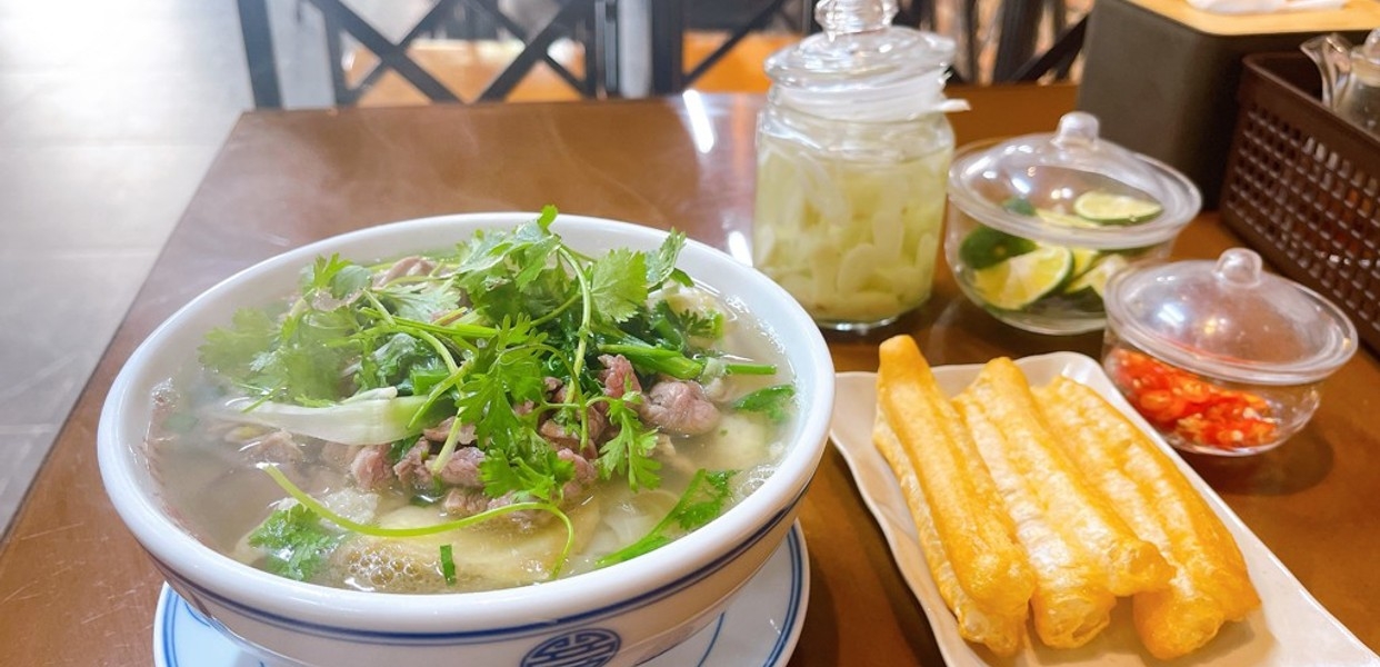 Bowl of beef pho topped with cilantro served with fried dough sticks and pickled garnishes on a wooden table
