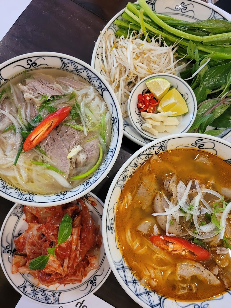 Two blue-rimmed bowls of pho, one clear beef and one spicy red, with kimchi, bean sprouts, herbs and lime