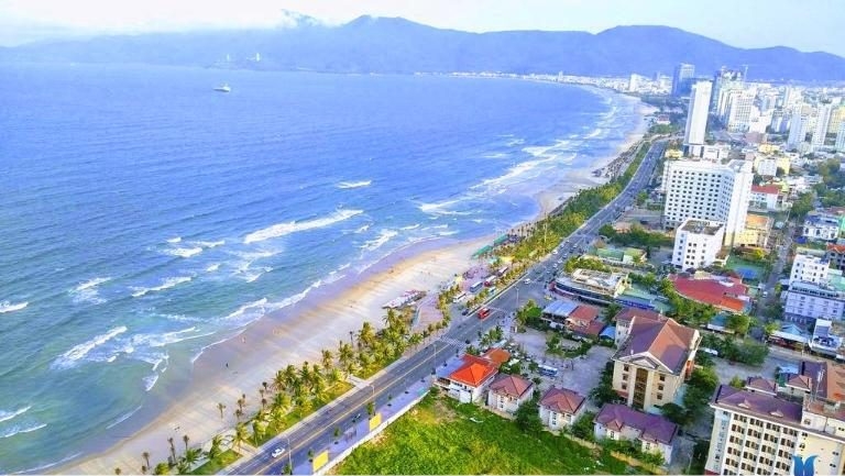 Aerial view of My Khe Beach in Da Nang, a long sandy shore beside a coastal road with city buildings and mountains