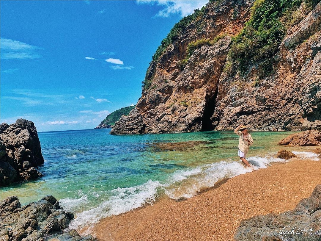 A person standing in shallow clear water at a small secluded cove with rugged cliffs and golden sand on Son Tra