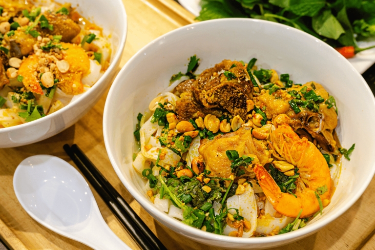 Bowls of Mi Quang with wide rice noodles, shrimp, peanuts, herbs and a ceramic spoon on wood