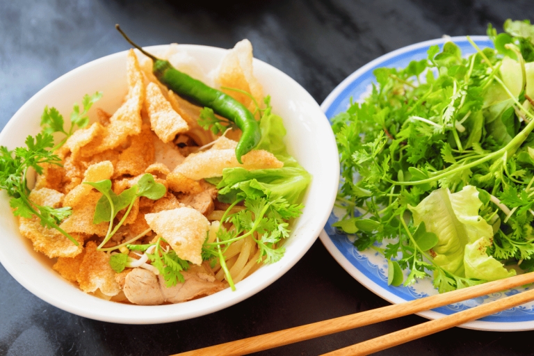 Bowl of Cao Lau noodles with pork, crispy crackers and herbs beside a plate of fresh greens and chopsticks