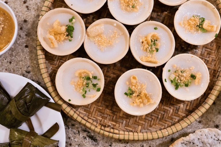 Small white Banh Beo rice cakes topped with crispy bits and scallions in a woven basket, leaf parcels nearby