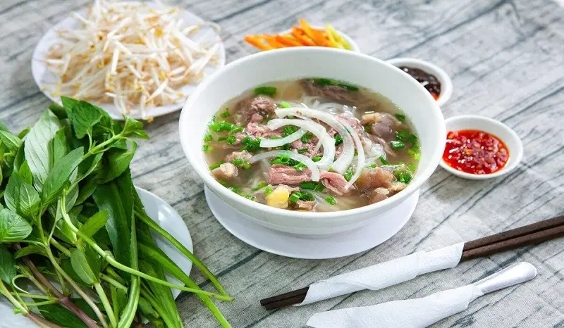 Bowl of beef Pho with sliced onion and herbs, served with bean sprouts, basil, chili sauce and chopsticks