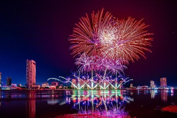 Pink and purple fireworks exploding over the riverside Da Nang skyline at night, mirrored on the water