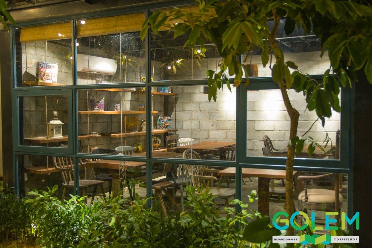 Golem Boardgames Coffeeshop seen through green-framed windows with wooden tables, brick walls and greenery