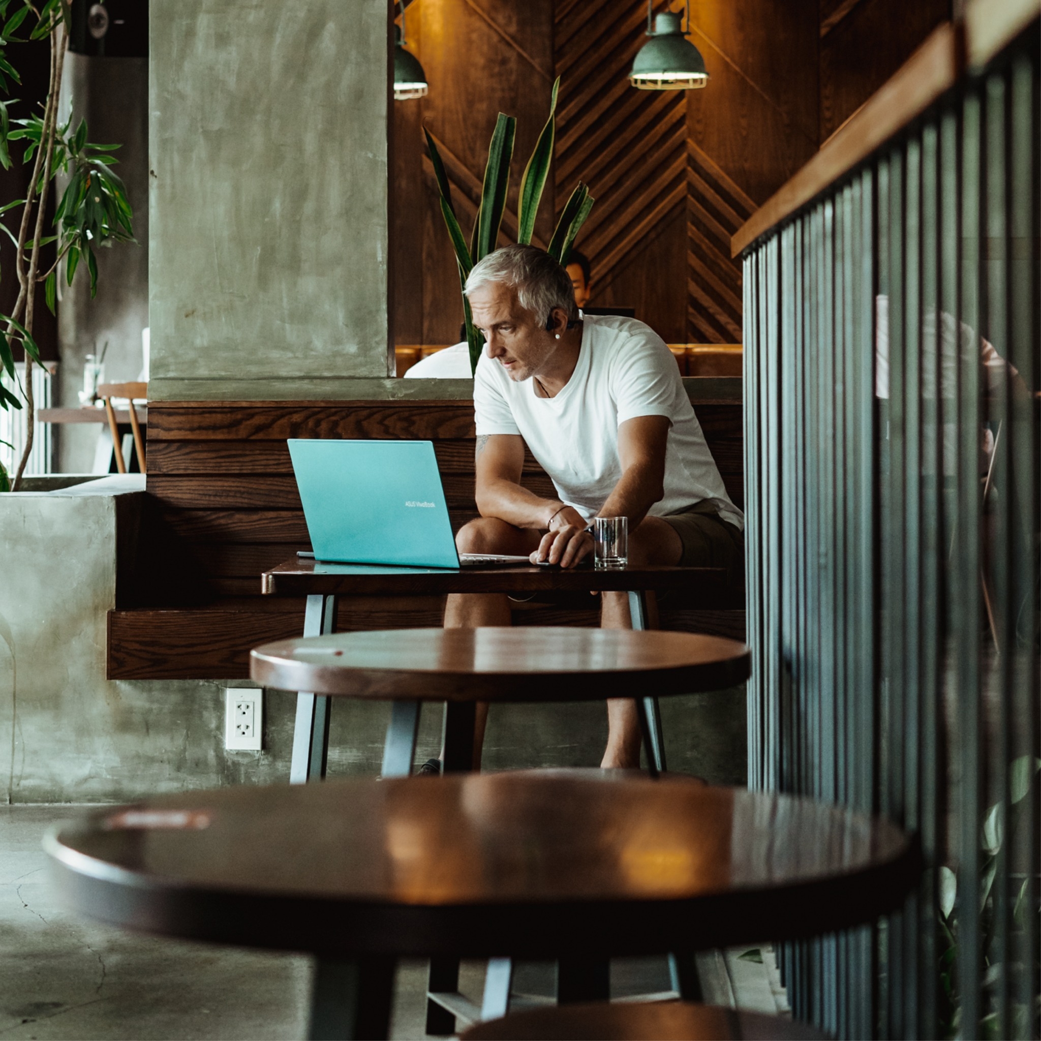 Man working on a laptop at a wooden table in a quiet café with plants and warm pendant lighting