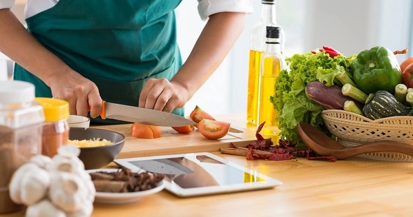 Person in a green apron slicing fresh tomatoes on a wooden board beside vegetables, herbs, olive oil and a tablet