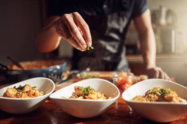 Hand sprinkling fresh herbs over three white bowls of steaming cooked shrimp in a warmly lit kitchen