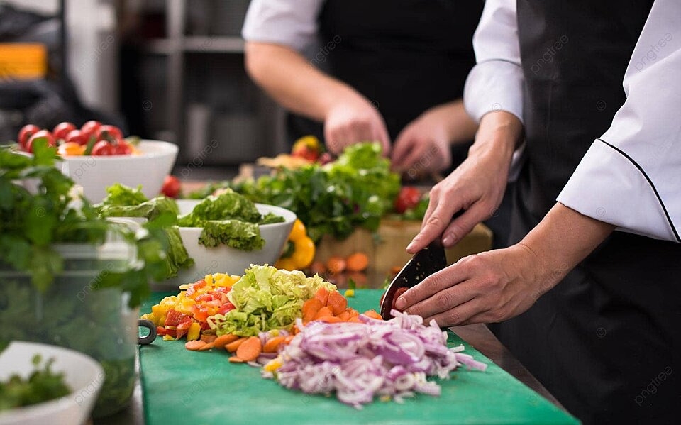 Two chefs in white jackets and black aprons chopping onions, carrots and peppers on a green cutting board