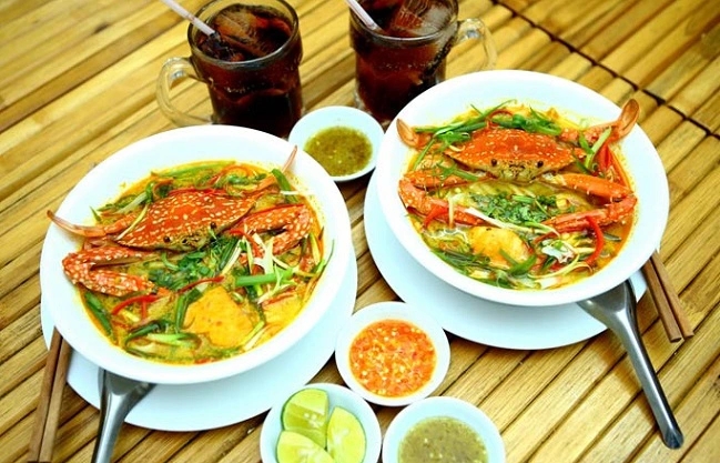 Two bowls of crab noodle soup with herbs served on a bamboo mat with iced drinks and dipping sauces
