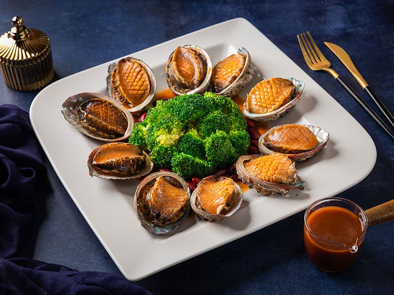 Cooked abalone in shells arranged around steamed broccoli on a white plate with dipping sauce