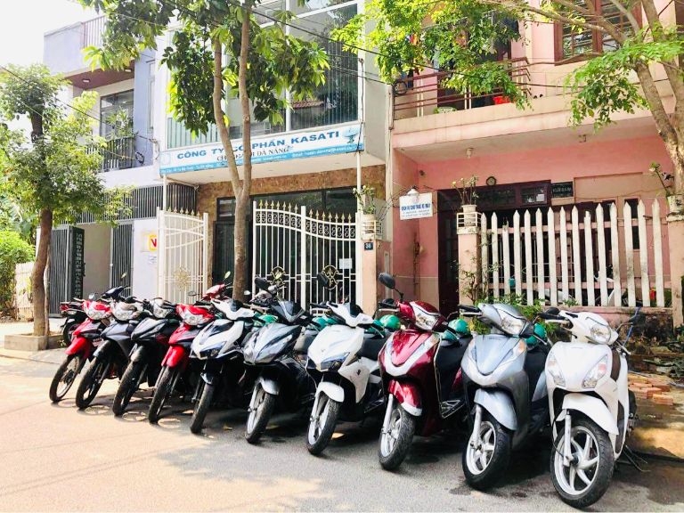 Row of rental scooters parked along a Da Nang street in front of a building with trees overhead