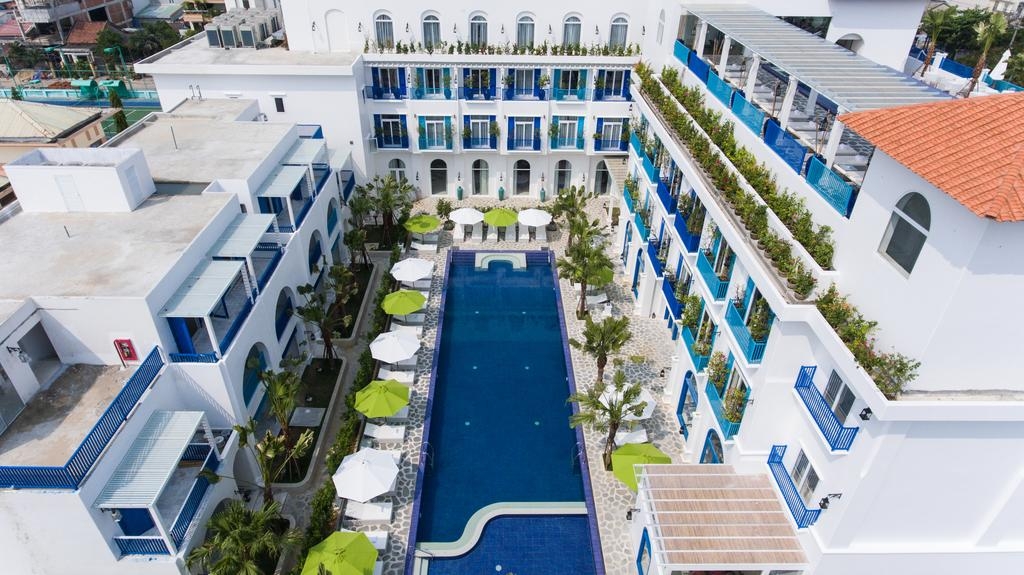 Aerial view of a white and blue Mediterranean-style building with a long central swimming pool, green umbrellas and balcony terraces