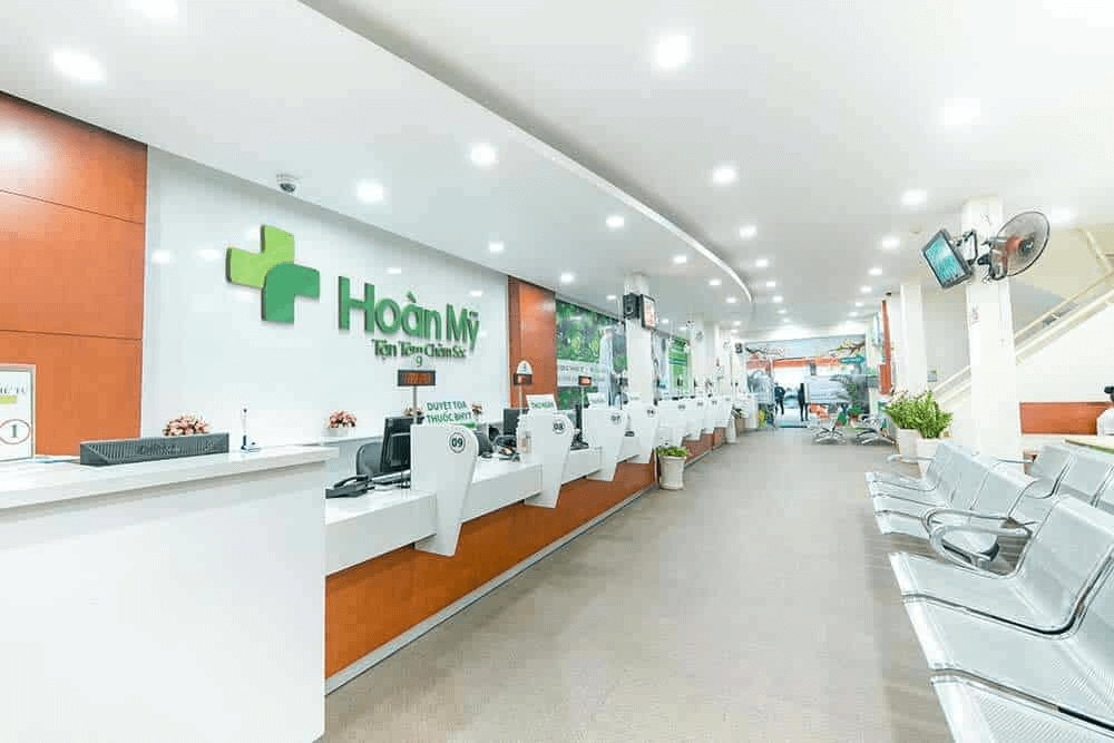 Bright lobby at Hoan My Hospital with green signage, white reception counters and rows of metal waiting chairs