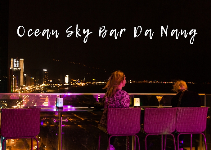 Two women sit at a rooftop bar at night overlooking a glowing city skyline, with pink and purple lighting along the railing