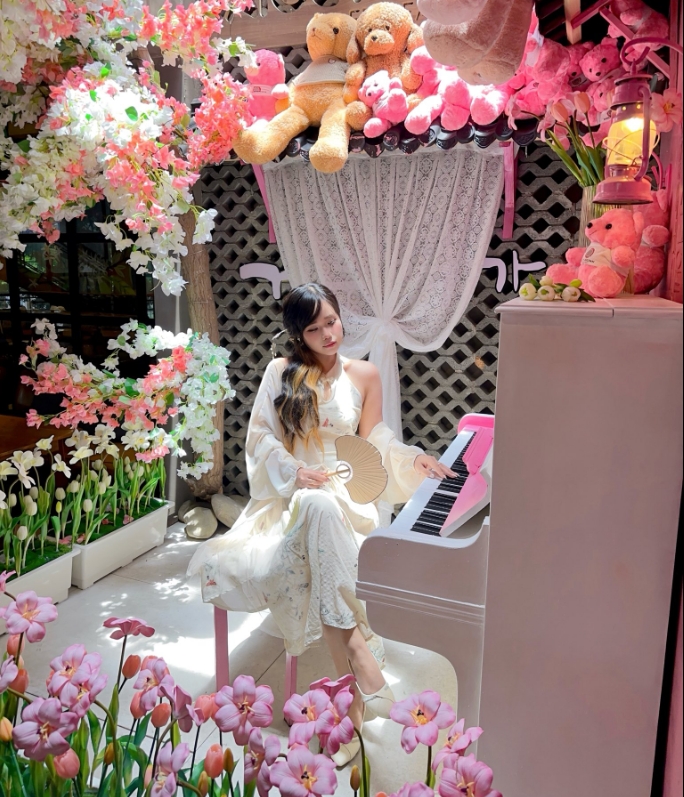 Woman in a white dress seated at a pink piano surrounded by cherry blossom branches, plush teddy bears and tulips