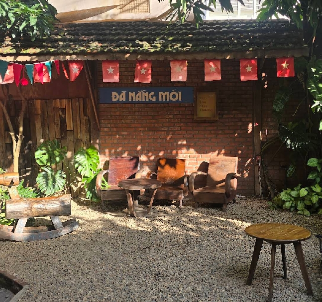 Rustic brick courtyard with wooden seating, a blue Da Nang Moi sign and small Vietnamese flag bunting strung overhead