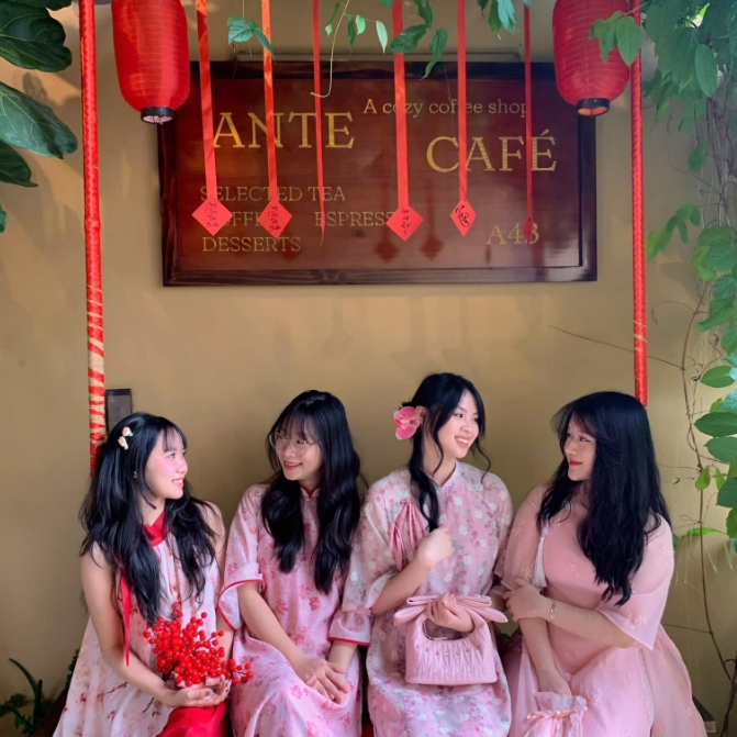 Four young women in pink ao dai seated on a bench under the wooden Ante Cafe sign with hanging red lanterns