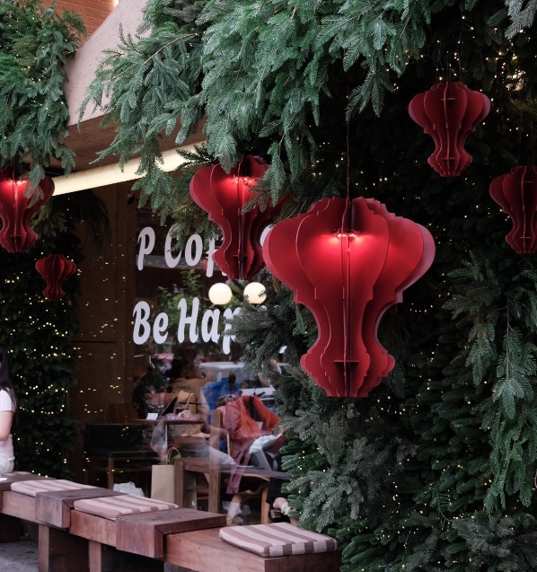 Cafe terrace lined with dense green pine foliage and large red paper lanterns beside a P Coffee Be Happy sign
