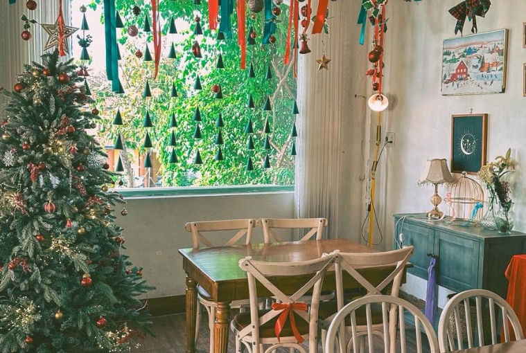 Cafe corner with a decorated Christmas tree, a window draped in tree-shaped felt garlands and ribbons, and wooden tables